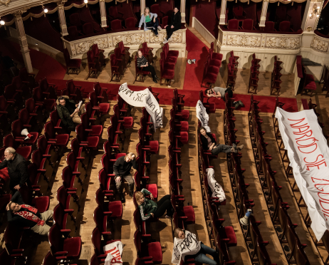 Wnętrze widowni dużej sceny teatru widziane z góry, z rzędami czerwonych foteli rozciągającymi się wzdłuż sali. Pomiędzy siedzeniami i na balkonach rozmieszczone są pojedyncze osoby, część z nich leży lub siedzi w swobodnych pozach. Wśród foteli rozłożone są białe transparenty z czerwonymi i czarnymi napisami.
